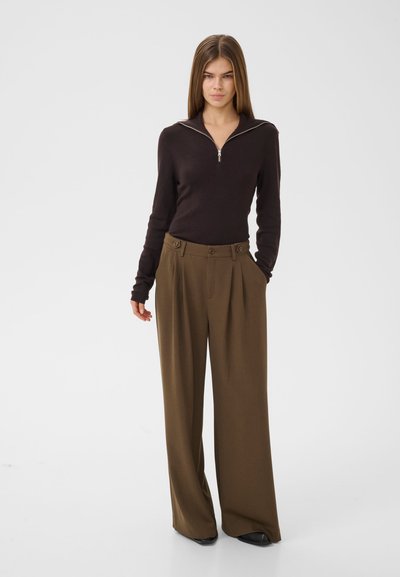 Femme aux cheveux longs et raides, portant un haut noir à manches longues avec fermeture éclair et un pantalon large marron ample, debout les mains dans les poches.