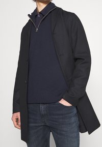 Abrigo negro con un diseño a medida, que presenta un cuello alto y botones frontales, superpuesto a un suéter azul marino oscuro y jeans, creando una silueta elegante.