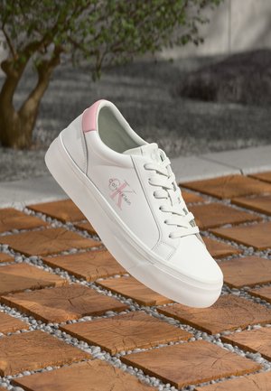 Baltos sportbačiai su rožiniu kulno akcentu, lygiu odiniu paviršiumi, plokščiomis raištelėmis ir šiek tiek pakeltu baltu padu; turi Calvin Klein logotipą.