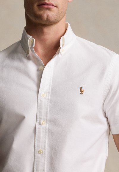 Camiseta branca de manga curta feita de algodão. Apresenta uma gola com botões e um pequeno logotipo bordado de um cavalo castanho no peito.