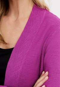 Purple cardigan with a ribbed detail along the edges, featuring a soft knit texture. Worn over a black top, displaying a V-neck design.