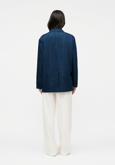 Γυναίκα με μακριά σκούρα μαλλιά που φοράει ένα σκούρο μπλε τζιν σακάκι και άνετο λευκό παντελόνι, στέκεται με την πλάτη σε απλό φόντο.