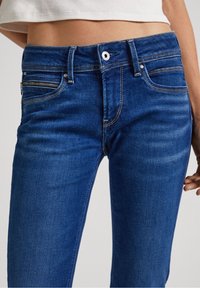 Blå denimjeans med en figurnära design, som har en dragkedjeficka, metallbeslag och kontrasterande sömmar vid midjan och framfickorna.