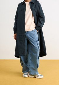 Czarna długa kurtka, jasnoróżowa bluza, luźne niebieskie spodnie cargo i białe sneakersy z siateczkowymi akcentami na żółtej podłodzie.