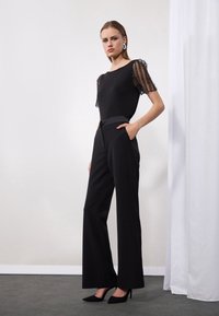 Top negru cu mâneci transparente și cu mărgele, asortat cu pantaloni negri cu talie înaltă. Modelul stă în tocuri negre ascuțite, pe un fundal deschis.