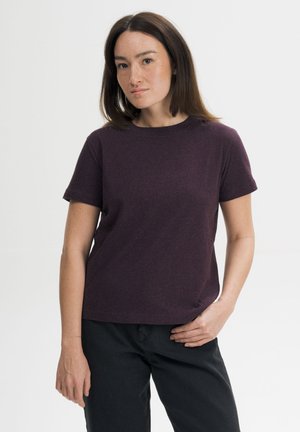 Vrouw met middellang bruin haar draagt een donkerpaars shirt met korte mouwen en zwarte broek, staand met één hand op haar heup.