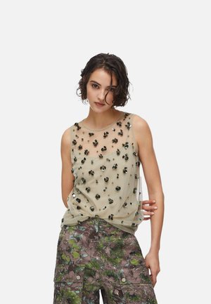 Giovane donna con capelli ricci corti che indossa una maglietta senza maniche trasparente decorata con paillettes nere e pantaloni cargo a fantasia.