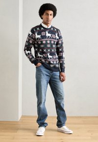 Sweater azul marinho com padrões festivos de rena e flocos de neve, punhos canelados e gola redonda, combinado com jeans azul claros e ténis brancos.