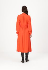 Mouwloze jurk tot aan de enkels in feloranje stof met een aansluitende taille, voorzien van een gladde textuur en eenvoudig ontwerp. Zwarte hoge laarzen zichtbaar.