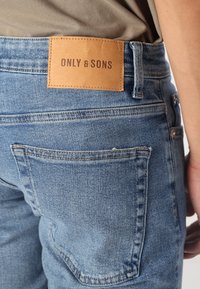 Modré džínsy z denimu s textúrovaným povrchom, s hnedou koženou nášivkou označenou "ONLY & SONS" na zadnom páse a štandardnými vreckami.