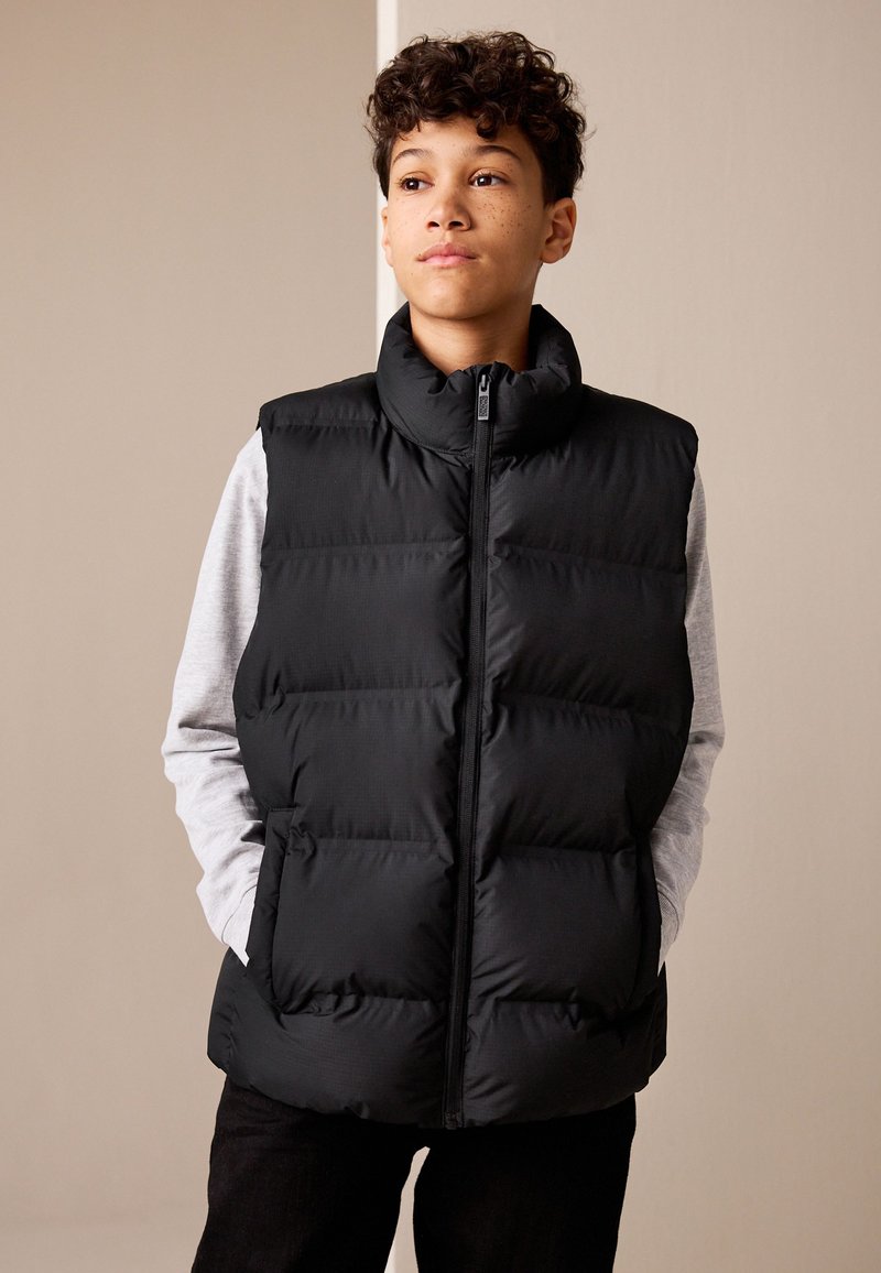 Teenager med krøllet hår og fregner iført sort dunvest over lys grå langærmet skjorte, hænderne i lommerne, kigger let mod højre.