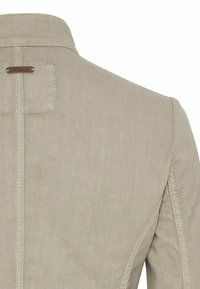 Beige tekstureret jakke set bagfra med lodret søm, firkantet lap og lille metalbrandmærke nær kraven på hvid baggrund.