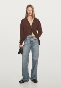 Hnědý pletený cardigan s černými knoflíky přes světle zelenou košili, v kombinaci s modrými džíny s vysokým pasem a černými klíny.