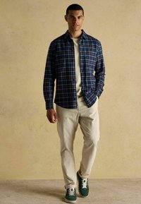 Vīriešu raibu kreklu jūras zilā, sarkanā un gaiši zilā krāsā, virsū valkājot bēša krāsas T-kreklu. Apvienots ar krēmkrāsas kordura biksēm un zaļiem sporta apaviem.