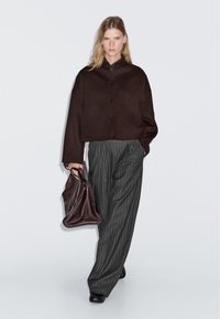 Donna che indossa una giacca buttonata marrone scuro, pantaloni larghi grigi a righe, scarpe nere, mentre tiene una grande borsa tessurizzata marrone scuro.