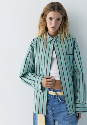 Junge Frau mit welligen blonden Haaren, die ein übergroßes, grün-gestreiftes, geknöpftes Hemd, ein weißes Crop-Top und blaue Jeans trägt, vor einem einfachen Hintergrund.