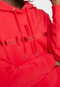Felpa rossa con coulisse e punte in metallo. Presenta il logo 'CK' in rilievo e la scritta 'CALVIN KLEIN JEANS' sul petto. Materiale in cotone.