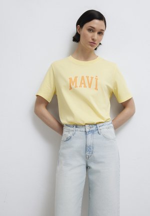 Woman with short dark hair wearing a yellow "MAVÌ" t-shirt and light blue high-waisted jeans, standing against a white wall.