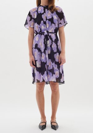 Femme portant une robe noire longueur genou avec de grands motifs floraux violets et blancs, manches courtes, taille nouée, et des ballerines noires pointues.