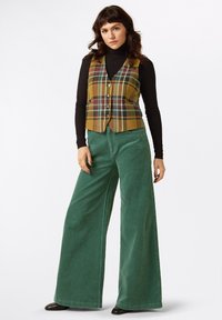 Kariertes Weste in Gelb, Grün und Rot mit fünf Knöpfen, kombiniert mit grünen, weit geschnittenen Cordhosen, die eine weiche Textur aufweisen. Schwarzer Rollkragenpullover darunter.