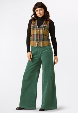 Gilet a quadri nei toni giallo, verde e rosso con cinque bottoni, abbinato a pantaloni larghi in corduroy verdi, caratterizzati da una consistenza morbida. Sotto una maglia a collo alto nera.