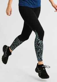 Gambe di una persona che corre, indossando leggings neri con un motivo a pois blu sui polpacci e scarpe da corsa nere, su uno sfondo chiaro.