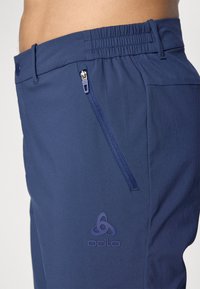 Navy blue athletic pants with an elastic waistband, a side zipper pocket, and a prominent embossed logo on the front. Smooth texture.
