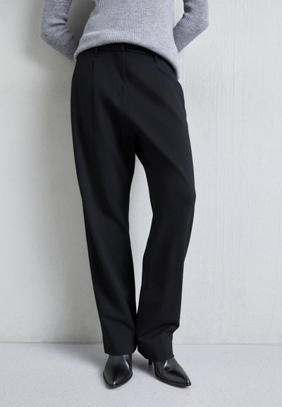 Pantalons noirs ajustés à taille haute, avec des plis et une coupe ample. Assortis à des bottes à talons aiguilles noires. La texture semble lisse.