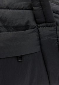 Gros plan sur un tissu noir matelassé avec des coutures surpiquées et une sangle épaisse, montrant la texture et un petit détail de boucle en métal.