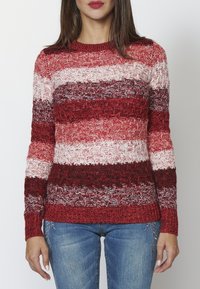 Sweter dziany w poziome czerwone, różowe i białe paski. Posiada teksturowany wzór oraz ściągacze przy dekolcie i rękawach. Połączony z dżinsami.