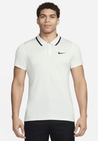 Jaunas vyras, vilkintis baltą trumpomis rankovėmis polo marškinėlį su juodu apykaklės apdailu ir juodu Nike logotipu krūtinėje, stovi prieš baltą foną.
