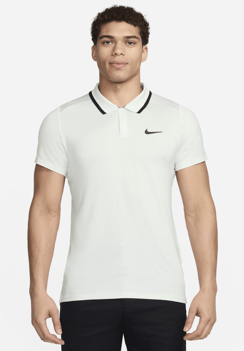 Mladý muž v bílé polokošili s krátkým rukávem, černým lemováním kolem límce a černým logem Nike na hrudi, stojící na bílém pozadí.