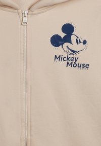 Sweat à zip beige avec un graphique de Mickey Mouse bleu marine orné de strass et d'une texture douce. Design simple avec capuche et fermeture éclair frontale.