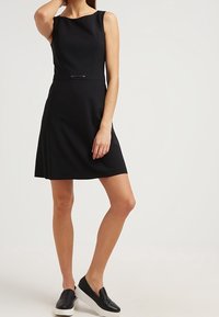 Vestido negro sin mangas con un cuerpo ajustado y falda acampanada; presenta un detalle plateado en la cintura. Combinado con zapatos negros de tipo slip-on.