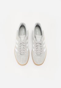 Szare zamszowe trampki z białymi paskami i gumowymi podeszwami. Posiadają zaokrąglony nosek i sznurowany design z oznaczoną językiem.