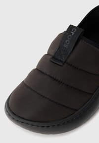 Svart slip-on-sko med en vadderad ovansida av tyg, med en texturerad gummisula och en framträdande svart etikett på det elastiska bandet.