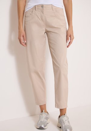 Pantalon beige droit avec fermeture par bouton et zip, porté avec des chaussures de sport argentées et un haut blanc.