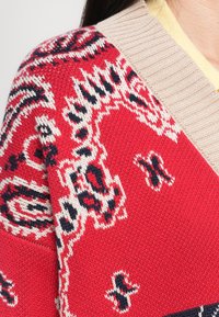 Gros plan sur un tricot rouge avec un col beige présentant des motifs géométriques et floraux complexes en noir et blanc sur l'épaule et la poitrine.