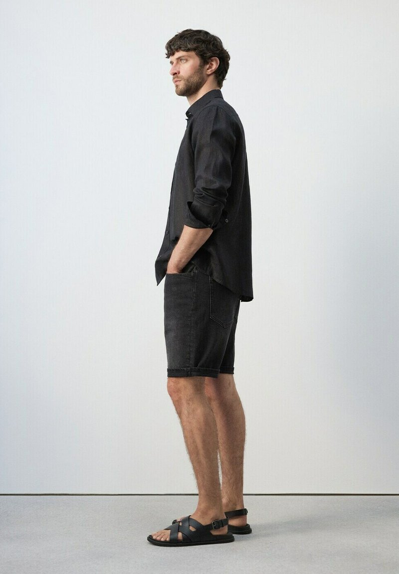 Homme debout de profil portant une chemise noire à boutons avec les manches retroussées, un short en jean noir et des sandales en cuir noire.