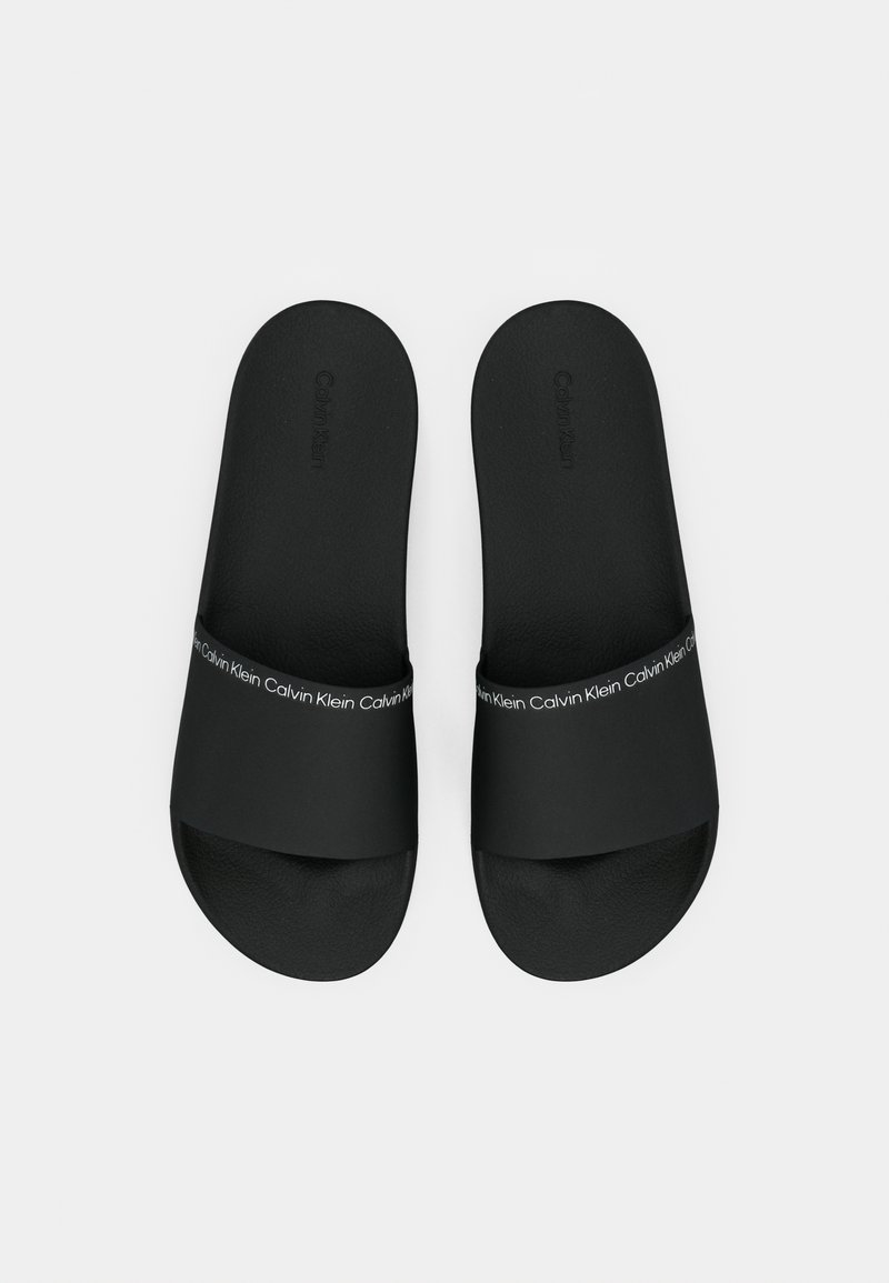 Sandales noires avec une large bandoulière, présentant une tige texturée et une semelle lisse. La bandoulière est décorée d'un logo "Calvin Klein" blanc répétitif.