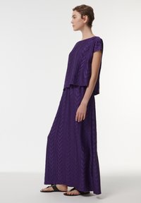 Paarse maxi-jurk met een gestructureerd, gelaagd ontwerp. Korte mouwen en een vloeiende silhouet, gecombineerd met zwarte platte sandalen.