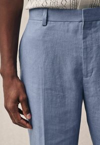 Pantalones de lino azul claro con una textura suave, con un frente plano y bolsillos laterales, mostrados con una mano cerca de la tela.