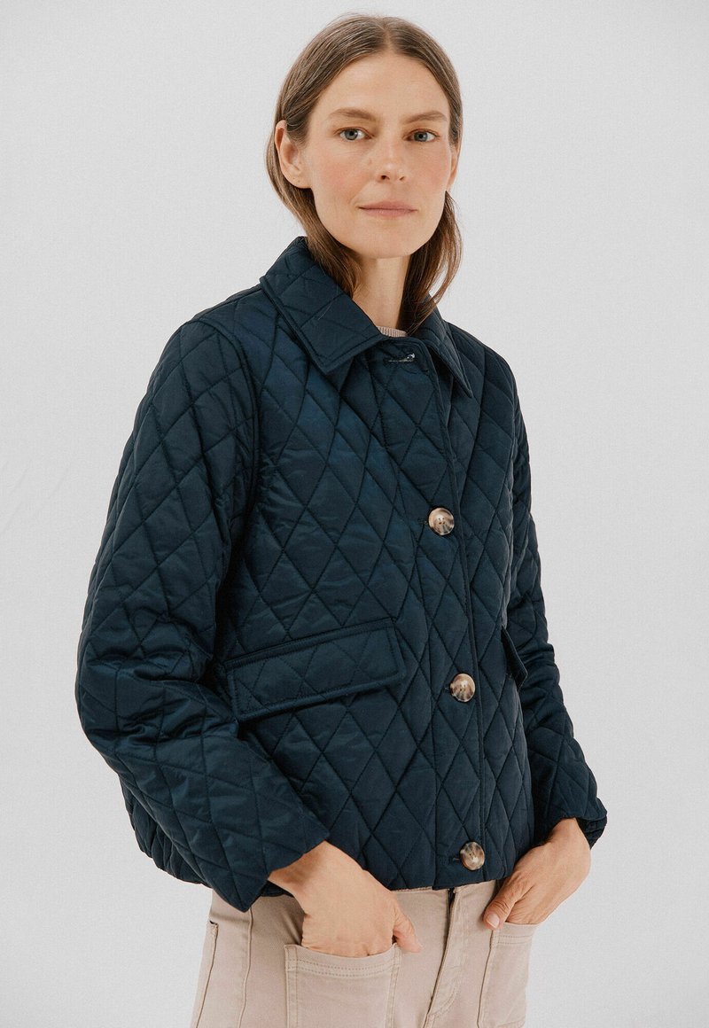 Marineblå quiltet jakke med diamantmønster, der har spidse kraver, store knapper og to sidelommer. Glat tekstur, afslappet design.