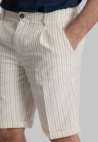 Pantaloni corti fino al ginocchio di un beige chiaro con sottili righe verticali. Presentano una chiusura con bottone, passanti per cintura e tasche laterali. Tessuto in cotone misto.