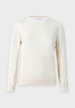 Kobieta z długimi blond włosami, nosząca okulary, biały sweter, ciemnozieloną spódnicę midi z rozcięciem z przodu oraz czarne botki, stojąca na białym tle.