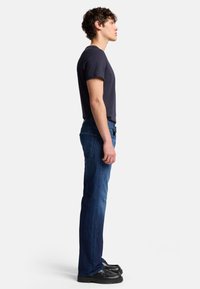 Slim-fit mörkblå jeans med rak ben, kombinerat med en åtsittande marinblå t-shirt och svarta skor, vilket visar en sidoprofil.