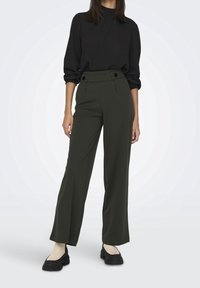Een model staat in een zwart hoge hals blouse en olijfgroene wijde broek, gecombineerd met zwarte plateau schoenen, tegen een lichte achtergrond.