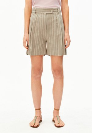 Frau trägt beige Shorts mit hohem Bund und vertikalen Streifen, kombiniert mit braunen flachen Sandalen, die mit Perlen verziert sind.