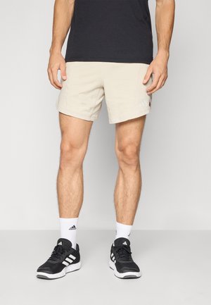 Shorts de sport beiges avec des coutures surfilées, une poche latérale et un logo Adidas, associés à des chaussures noires et des chaussettes blanches.