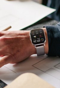 Montre intelligente avec un écran tactile carré, bracelet en maille métallique, affichant l'heure 09h26 et les détails météorologiques sur un fond sombre.